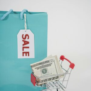 Miniature shopping cart with cash next to a sale-tagged bag, illustrating shopping and savings.
